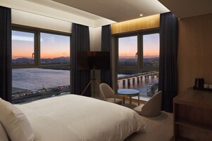 Habitación de hotel con una cama grande, un escritorio, una silla y un televisor. Amplias ventanas ofrecen vistas a la ciudad y un puente.