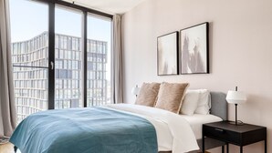 Un dormitorio moderno con un ventanal grande, una cama con una manta azul, dos almohadas, una mesita de noche con una lámpara y dos cuadros enmarcados en la pared.