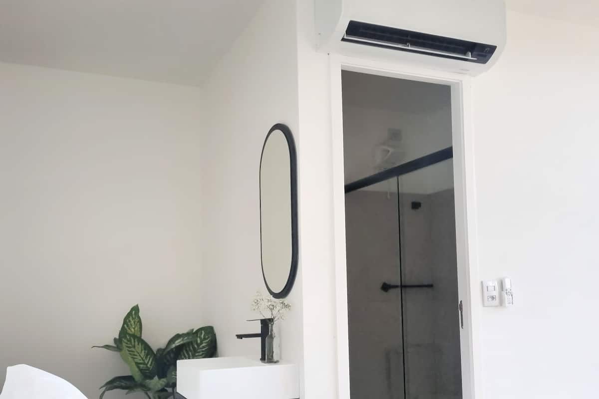 A hotel room with a bed, a white bedside table with a mirror, a potted plant, a shower area, and a wall-mounted air conditioning unit.