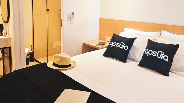 Hotelkamer met een bed, twee zwarte kussens met de tekst 'capsula', een laptop, een hoed en een spiegel aan de muur.
