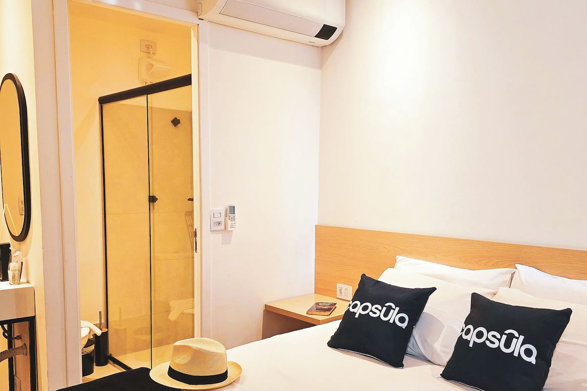 A hotel room with a bed, two black pillows with 'capsula' text, a laptop, a hat, and a mirror on the wall.