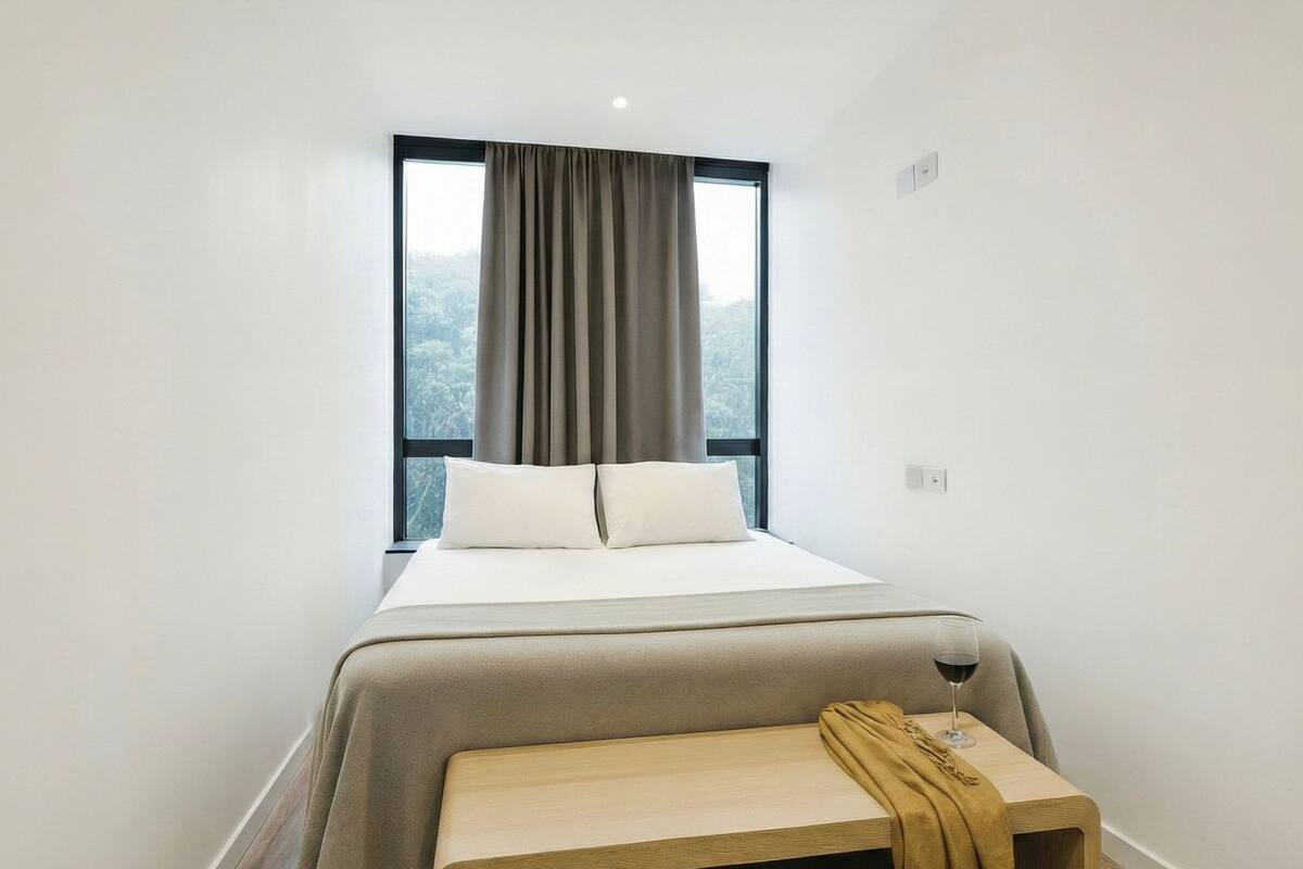 A minimalist hotel room with a large window, a bed with white linens, a wooden bench, and a glass of wine.