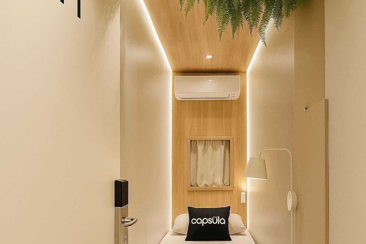 A compact hotel room with a bed, a bedside lamp, an air conditioning unit, and a plant hanging from the ceiling.
