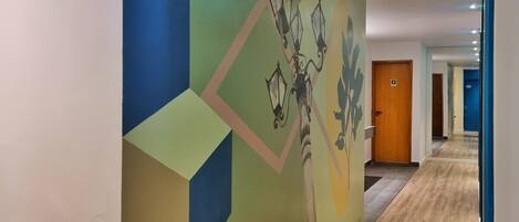 Un couloir orné d’une fresque géométrique, doté d’un plancher en bois et d’une porte sur laquelle figure un numéro.