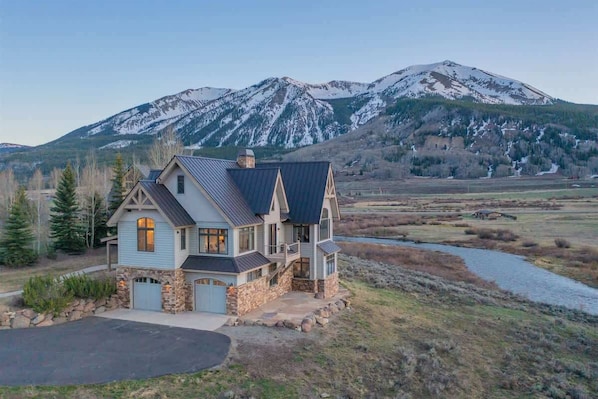 A house with a stone foundation, a blue exterior, and a metal roof, surrounded by a natural landscape with a mountain in the background.