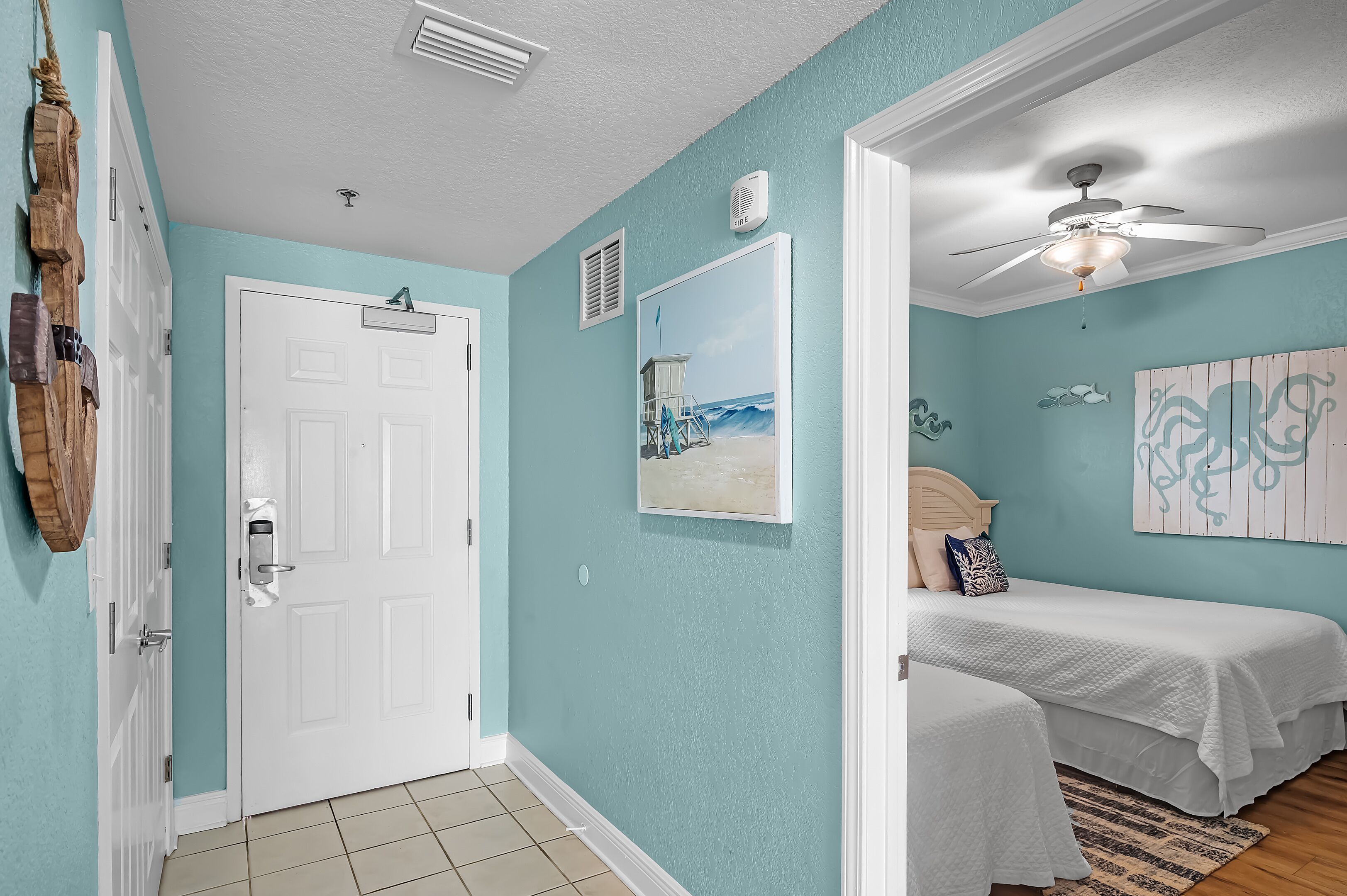 A room with teal walls, a white door, and a ceiling fan. There are two beds with white bedding and a painting of a beach scene.