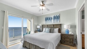 A bedroom with a large bed, a ceiling fan, a nightstand with a lamp, a dresser, and a view of the ocean through a sliding door. - Crystal Shores West 701 Corner Condo Wraparound Balcony | Indoor & Outdoor Pools | Beach Getaways (Gulf Shores)