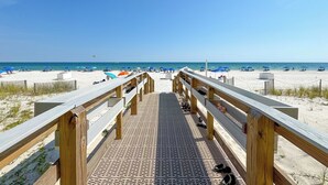 A wooden boardwalk leading to a beach with umbrellas and people. - Crystal Shores West 701 Corner Condo Wraparound Balcony | Indoor & Outdoor Pools | Beach Getaways (Gulf Shores)