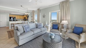 A spacious living room with a gray sofa, striped armchair, and a round coffee table. A ceiling fan and a chandelier are visible. - Crystal Shores West 701 Corner Condo Wraparound Balcony | Indoor & Outdoor Pools | Beach Getaways (Gulf Shores)