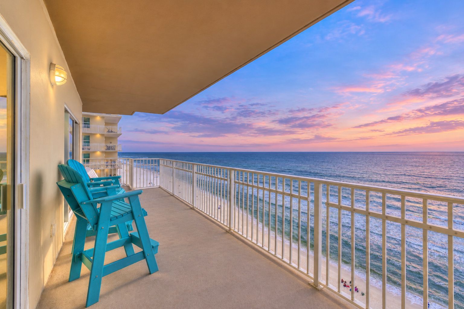 A balcony with a view of the ocean at sunset, a railing, and two chairs.