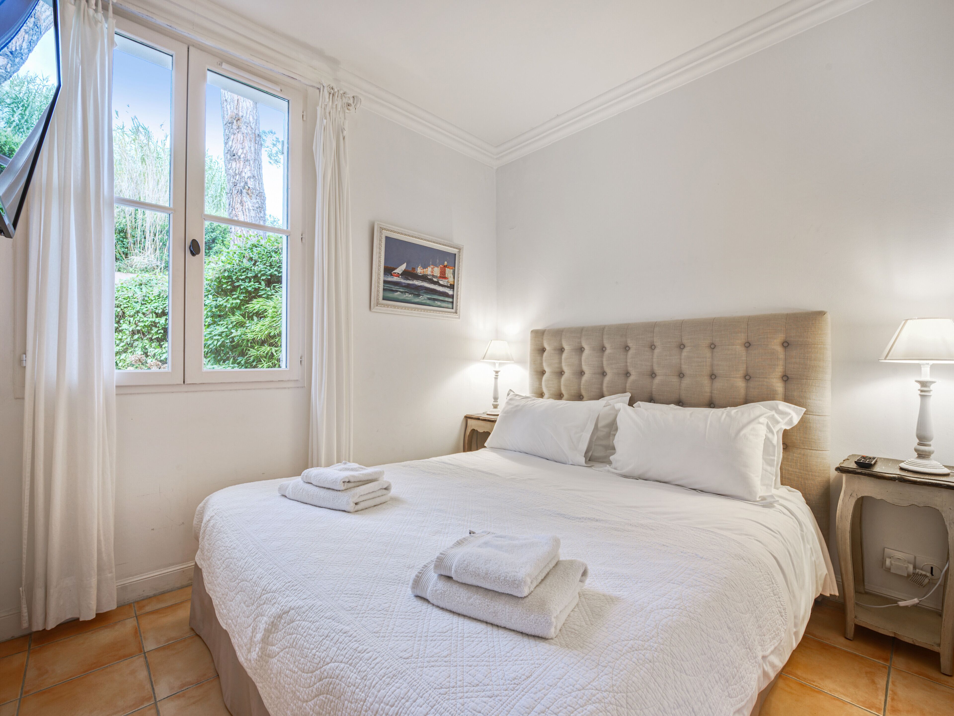 Ein ordentlich bezogenes Bett mit weißen Leinentüchern und Handtüchern, ein großes Fenster mit Vorhängen, ein gerahmtes Bild an der Wand und ein Nachttisch mit einer Lampe.