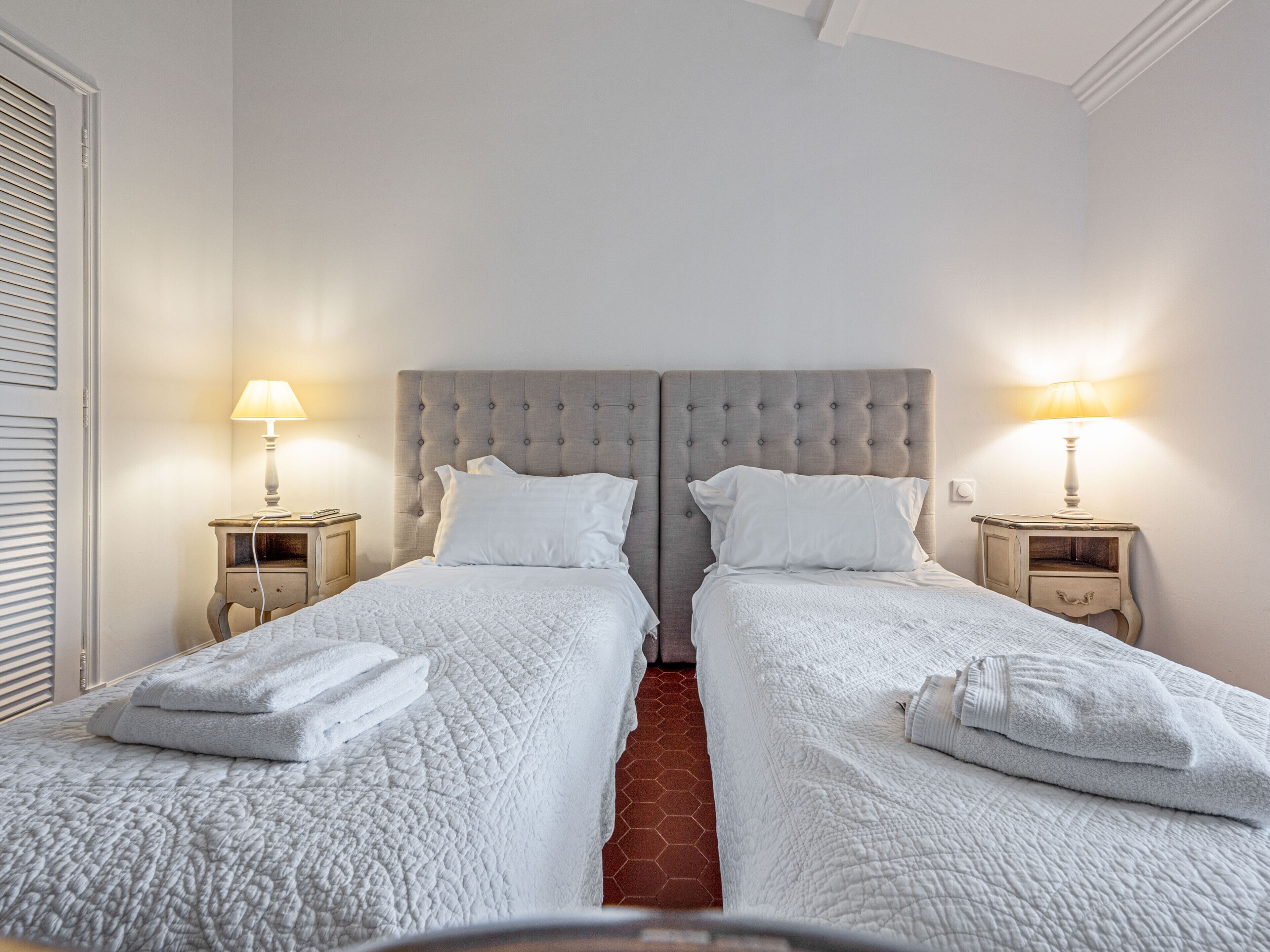 Une chambre double avec deux lits, chacun recouvert d’une courtepointe blanche et agrémenté d’oreillers assortis. De chaque côté des lits, on trouve des tables de chevet avec des lampes.