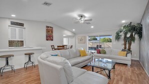 Una sala de estar moderna con un sofá blanco, una mesa de centro de vidrio y un ventilador de techo. Hay dos taburetes de madera y una barra blanca con un letrero que indica cava de vinos.
