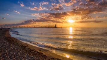 Una playa con un muelle que se adentra en el agua, un faro a lo lejos, y el sol poniéndose en el horizonte.