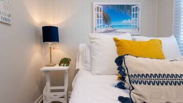 Un dormitorio con una cama, una mesita de noche con una lámpara, una pequeña mesa con una planta y un cuadro de una playa.