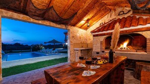 Un espace extérieur aménagé pour les repas, avec une cheminée en pierre, une piscine et une vue sur le ciel du soir.