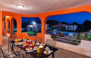 Ein Essbereich im Freien mit einem für das Frühstück gedeckten Tisch, mit Blick auf einen Pool und ein Steinhaus.