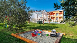 Un espace de jeux sablonneux avec des jouets, une clôture en bois et un bâtiment aux arches en arrière-plan.