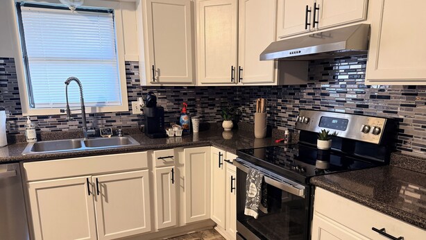 A modern kitchen with white cabinets, a black countertop, a stainless steel range hood, and a tiled backsplash.