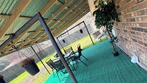 An outdoor patio with a green artificial turf, a table with chairs, hanging lights, and a brick wall.