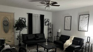 A living room with a black sofa, a coffee table, a desk with a computer, and a plant.