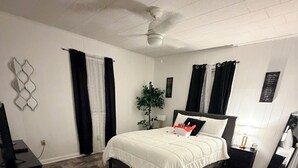 A bedroom with a bed, a desk, a plant, and a ceiling fan.