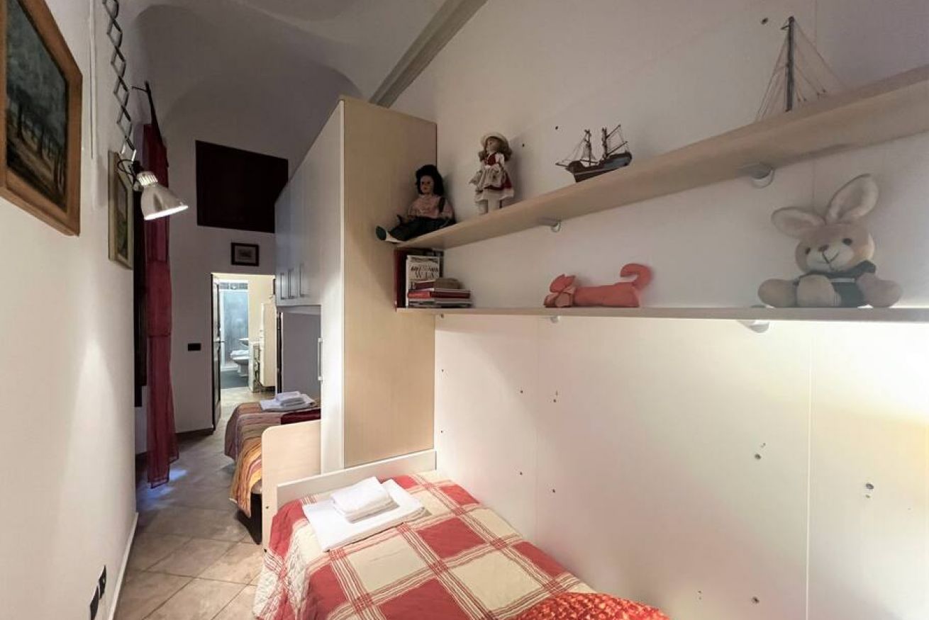 A narrow hallway leading to a bedroom with a bed, a small table, and a shelf with various items.