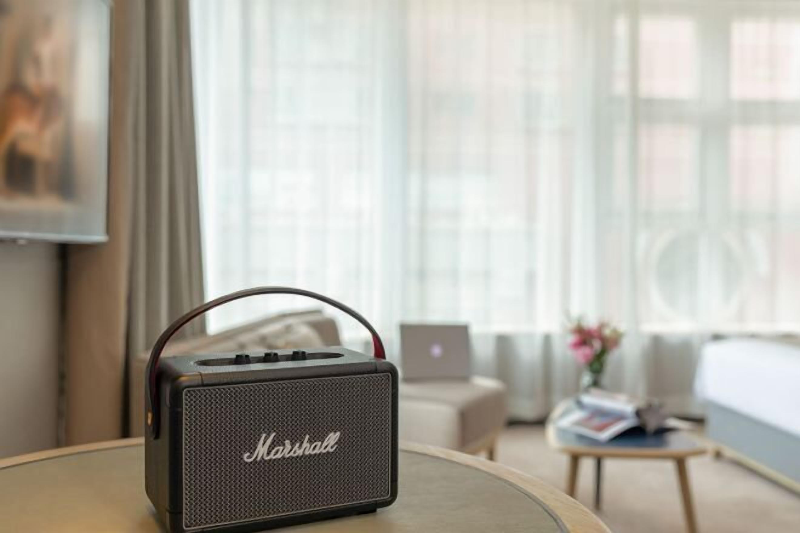 A Marshall portable speaker on a table in a hotel room with a laptop, a sofa, and a coffee table.