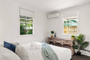 A bedroom with two large windows, a wooden dresser, a potted plant, and a bed with white linens.