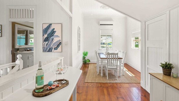 A bright, white interior with a wooden floor, a dining area with white chairs, a kitchen with white cabinets, and a large window providing natural light. - Family Home in Balmoral - Family Stays (Balmoral)