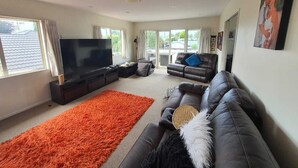A modern living room with a flat-screen TV, leather sofas, a large orange rug, and a balcony with a view. - Spacious 3 brm home for families to enjoy in Tauranga ga (Tauranga)