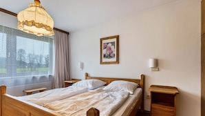 Un lit double avec une tête de lit et un pied de lit en bois, deux tables de chevet, une petite table d’appoint, une fenêtre avec des rideaux légers, un cadre photo fixé au mur et un luminaire suspendu.