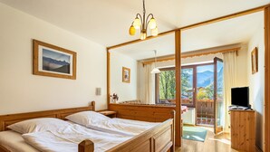 Een houten bed met witte lakens, een nachtkastje van hout, een ingelijst foto aan de muur, een televisie op een houten kast, en een balkon met uitzicht op bergen.