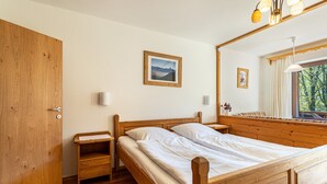 Een houten bed met nachtkastje, een houten garderobe, een spiegel en een raam met uitzicht op bomen.