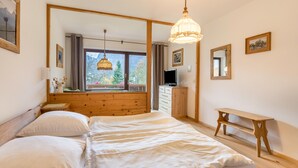 Quarto com cama grande, cabeceira de madeira, mesa de cabeceira, um banquinho, televisão e janela com vista para árvores.