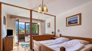Een bed met houten frame en witte lakens, een televisie op een houten dressoir, een nachtkastje van hout, een ingelijst foto aan de muur, en uitzicht op bergen door een raam.