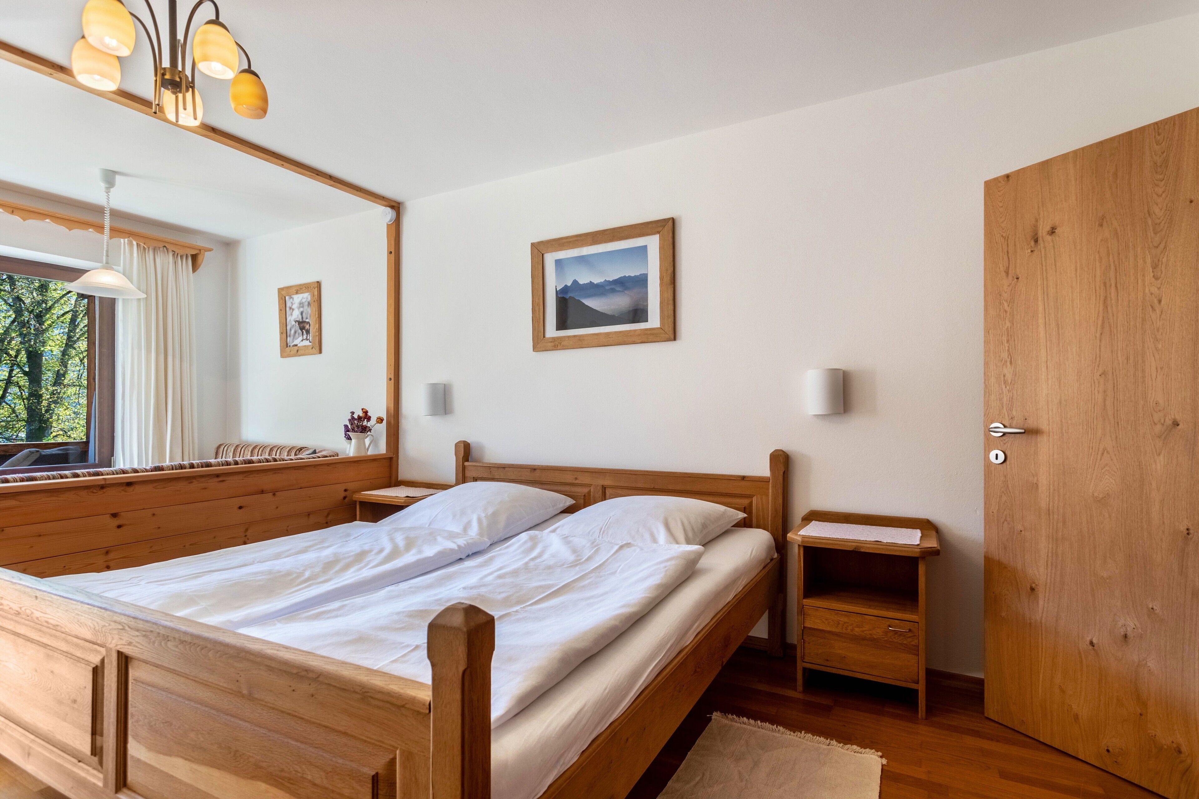 Een houten bed met witte lakens, een nachtkastje, een spiegel, een ingelijst beeld en een houten garderobe.