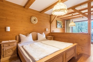 Un dormitorio con paneles de madera, una cama doble, mesitas de noche, una araña y una decoración en la pared. Se aprecia una vista por la ventana.