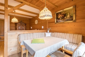 Interior de una cabaña de madera con una mesa de comedor, asientos tipo banco, una lámpara colgante, un armario y un cuadro enmarcado.