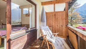 A wooden deck with a cushioned chair, a bed with a purple bedspread, and a view of mountains.
