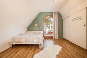 Een slaapkamer met een houten vloer, een bed met een witte spreidek, een wit vloerkleed, een zeshoekige lamp en een deur met een citaat.