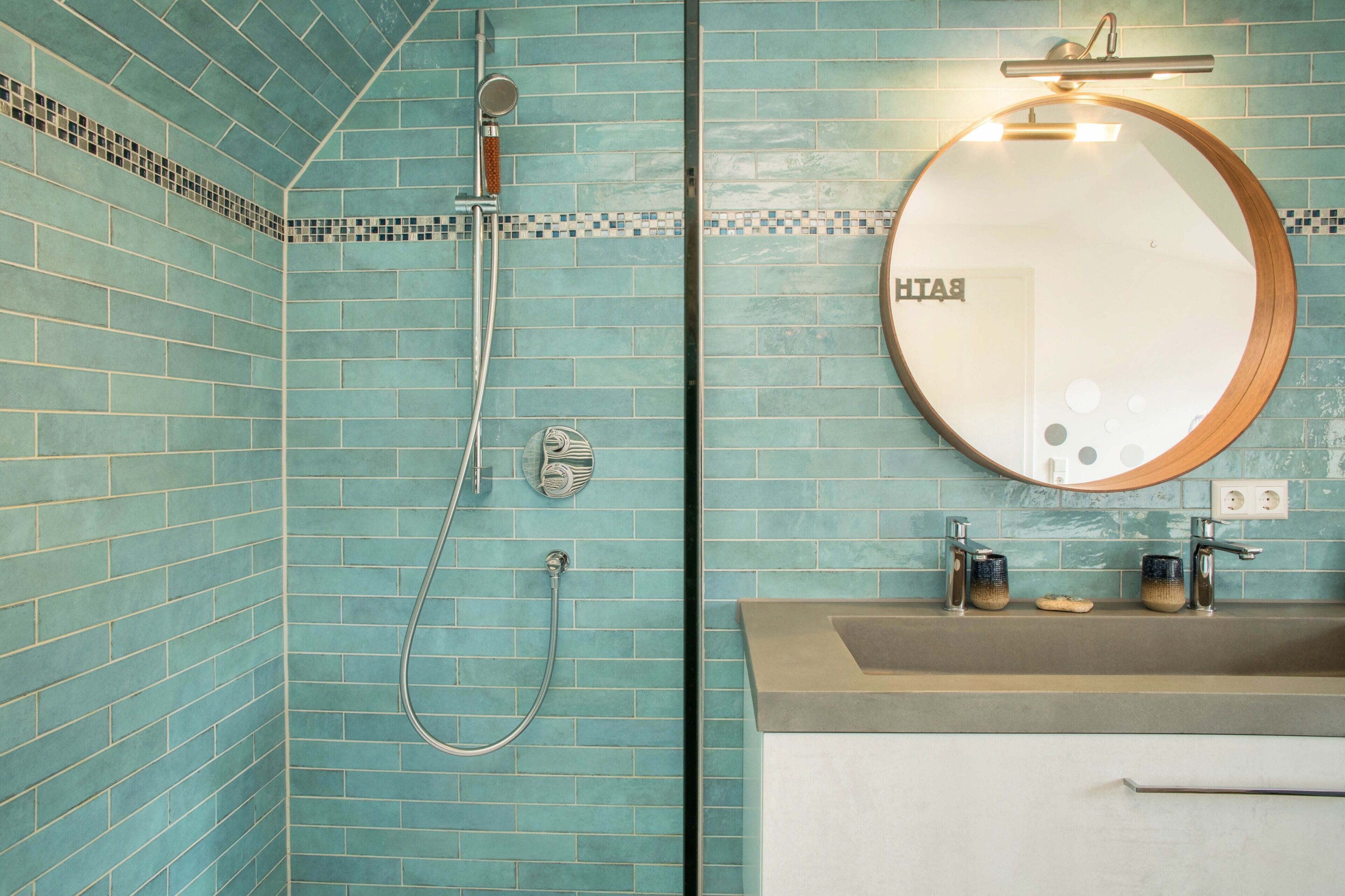 A bathroom with turquoise tiles, a round mirror, a shower, and a sink with two faucets.