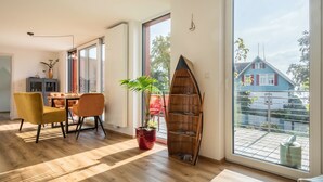 Een moderne woonkamer met een houten vloer, een eettafel met stoelen, een potplant, een boekenkast en een groot raam met uitzicht op een huis.