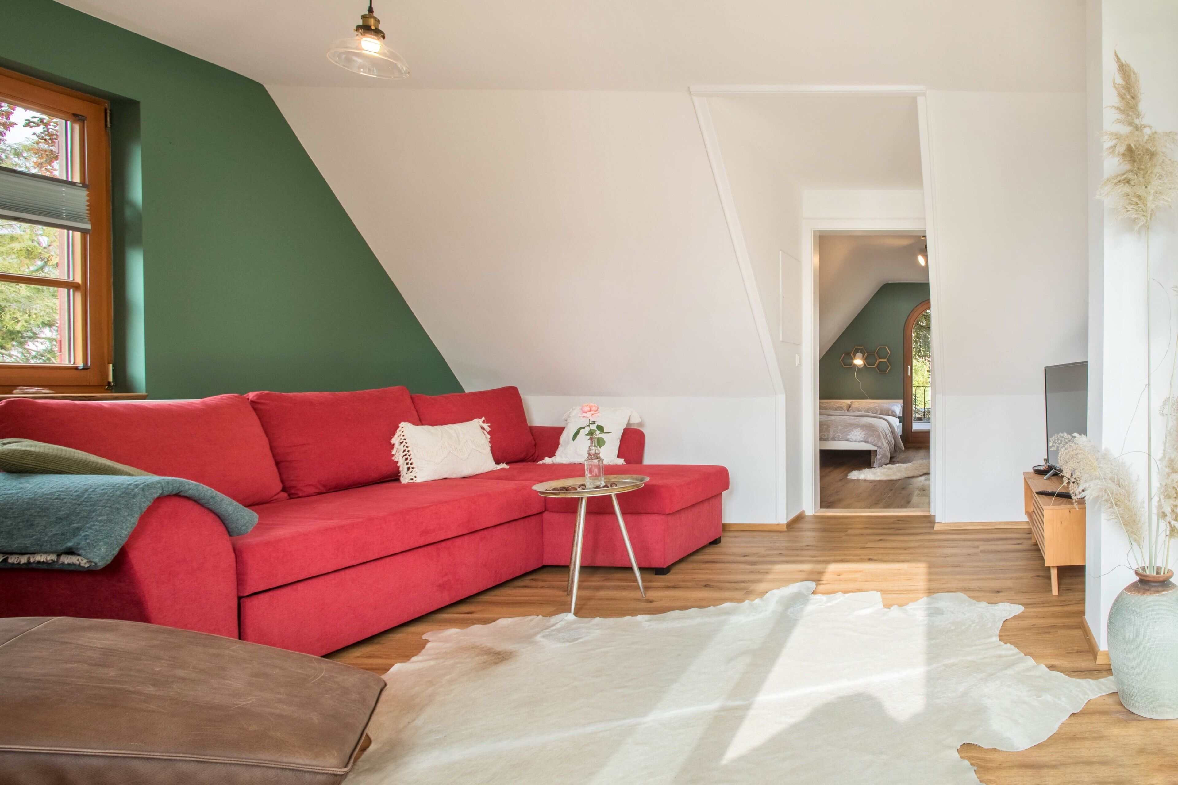 Een rode bank, een klein rond tafeltje, een wit vloerkleed en een houten vloer in een kamer met groene wanden en een schuine plafond.