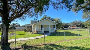 A single-story house with a front porch, surrounded by a fenced yard and a large tree.