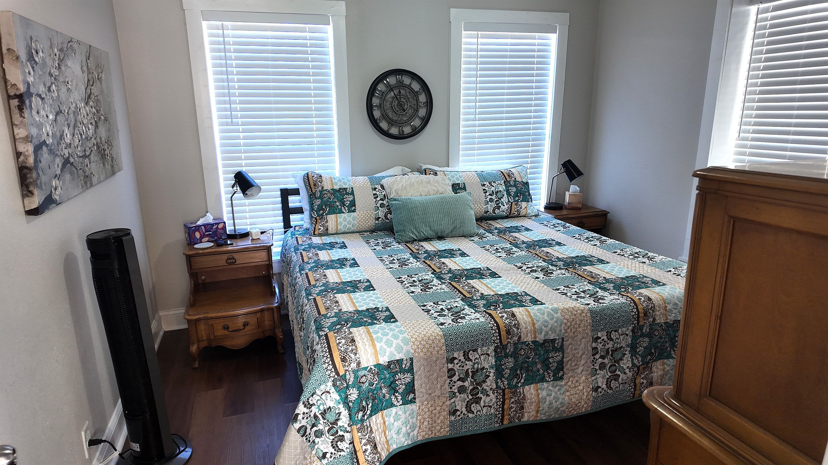 A bedroom with a bed, two bedside tables, a wall clock, and a window with blinds.