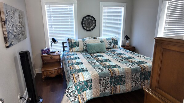 A bedroom with a bed, two bedside tables, a wall clock, and a window with blinds.