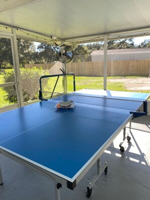 Mesa de ping pong al aire libre con red, bajo una zona techada con ventilador de techo.