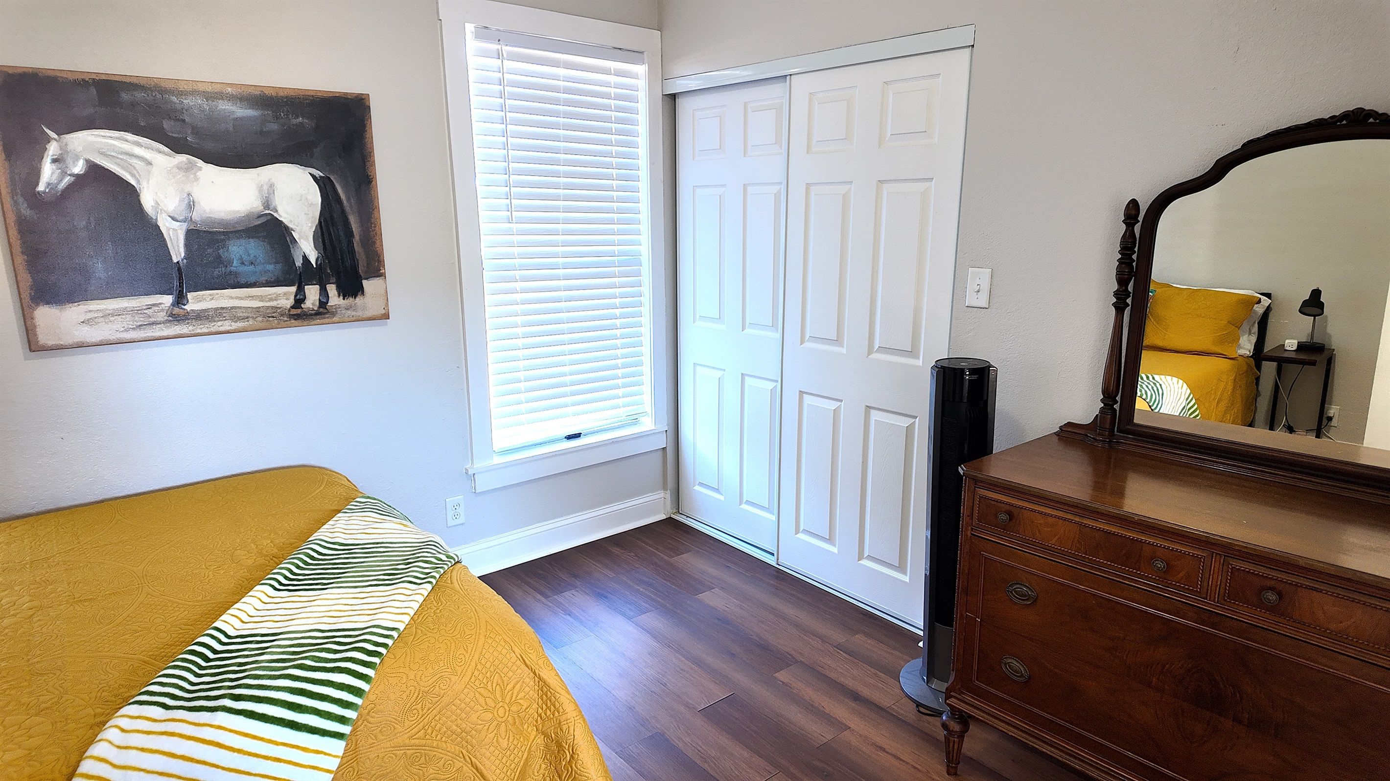 A bedroom with a bed, a dresser, a mirror, a painting of a horse, and a closed door.