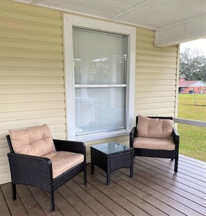 A porch with two wicker chairs and a small table, a window with blinds, and a view of a grassy area and a building in the background.